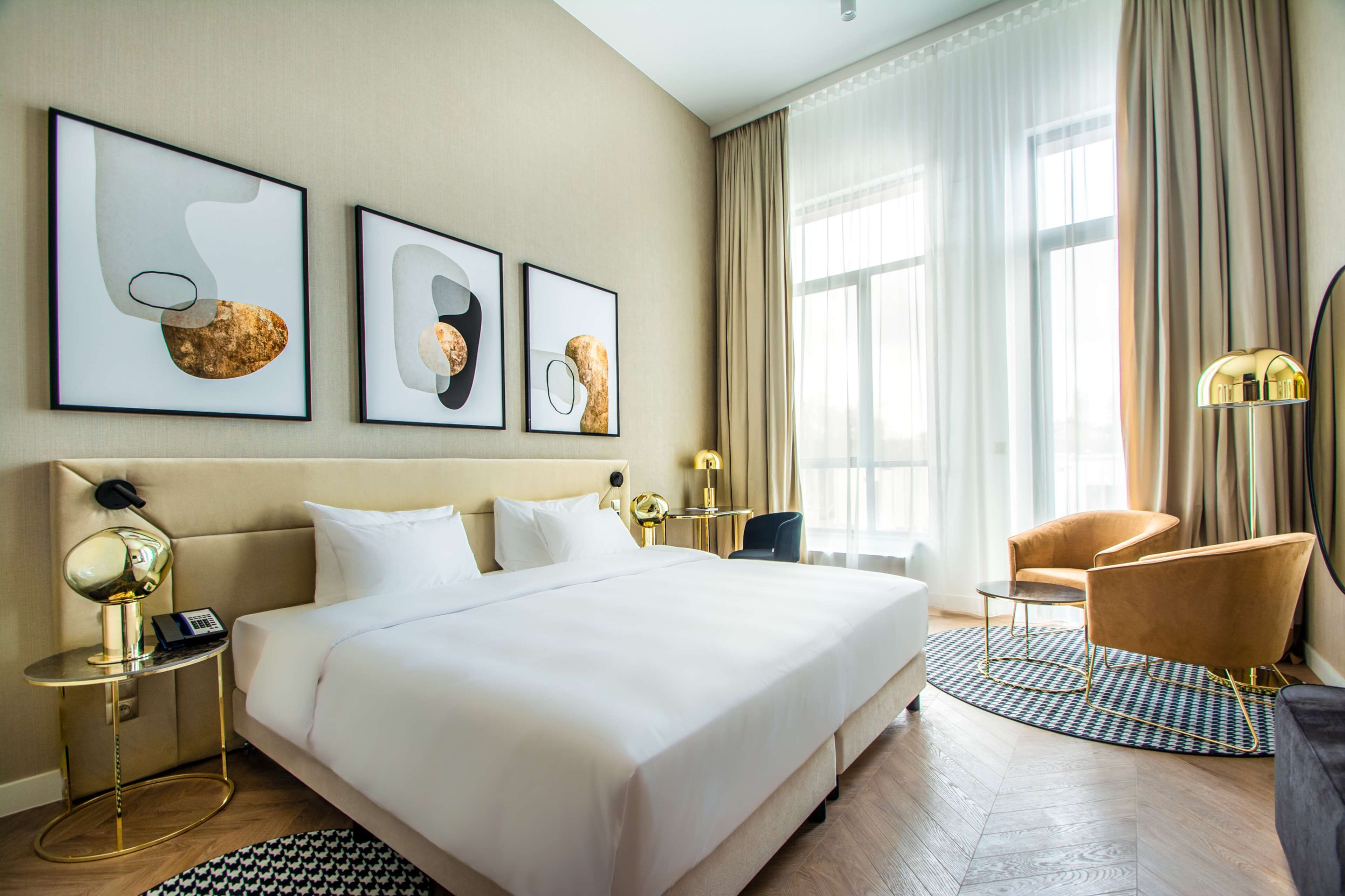 A hotel room with a large bed, a desk, a chair, and three framed artworks on the wall.