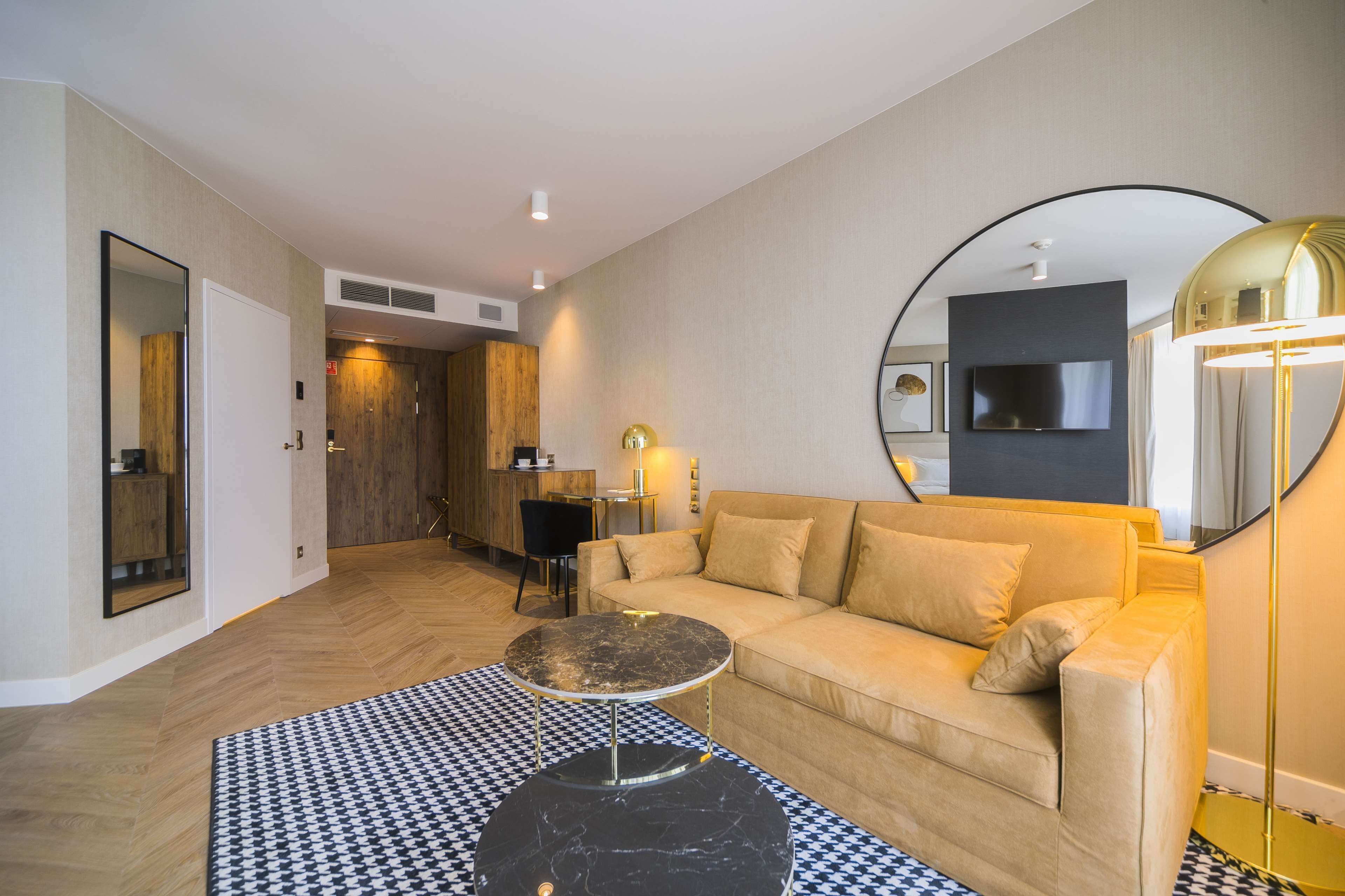 A modern living room with a sofa, a round coffee table, a checkered rug, a large mirror, and a television mounted on the wall.