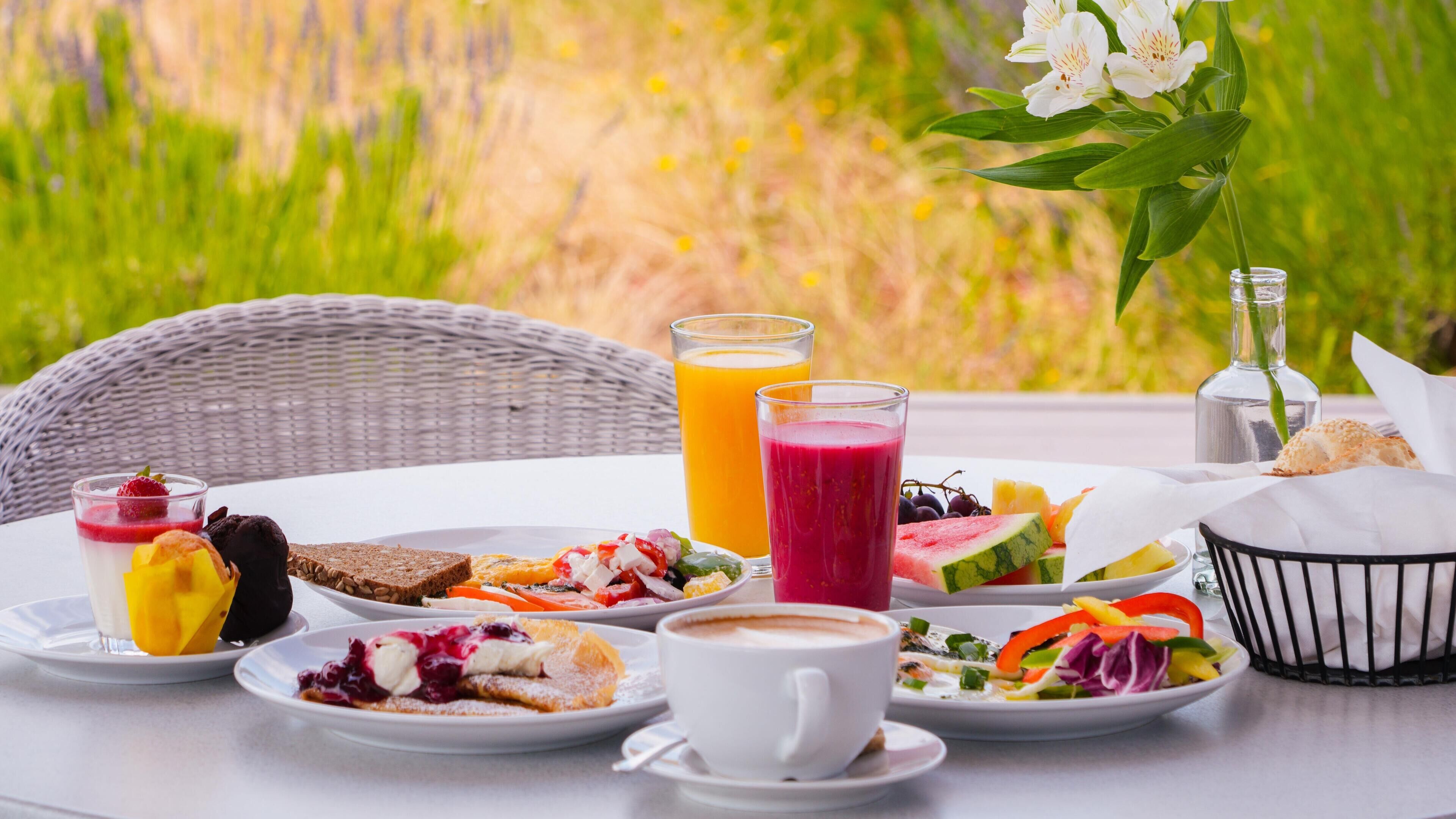 Un desayuno con una silla de mimbre, una mesa con platos de comida, vasos de zumo de naranja, un batido, una taza de café y una cesta de pan.