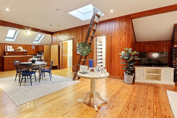 A spacious living area with a wooden floor, a dining table with chairs, a ladder, a TV, and a large plant. - Heated Pool Access | 5*Clean | Flex Cancelation (Savannah)