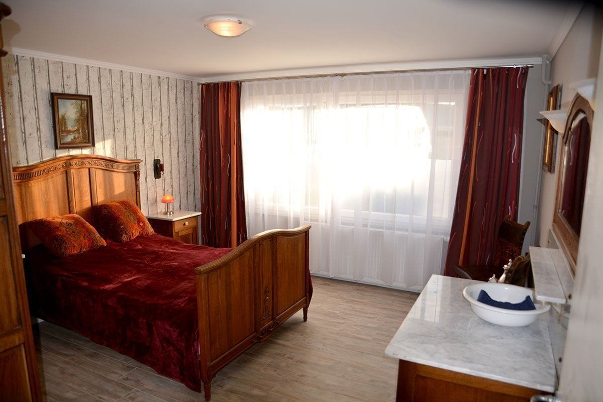 A bedroom with a wooden bed, a nightstand, a marble sink, and a large window with curtains.