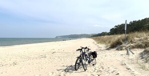 Uma praia arenosa com bicicletas estacionadas perto da beira-mar, céu claro e área arborizada ao fundo.