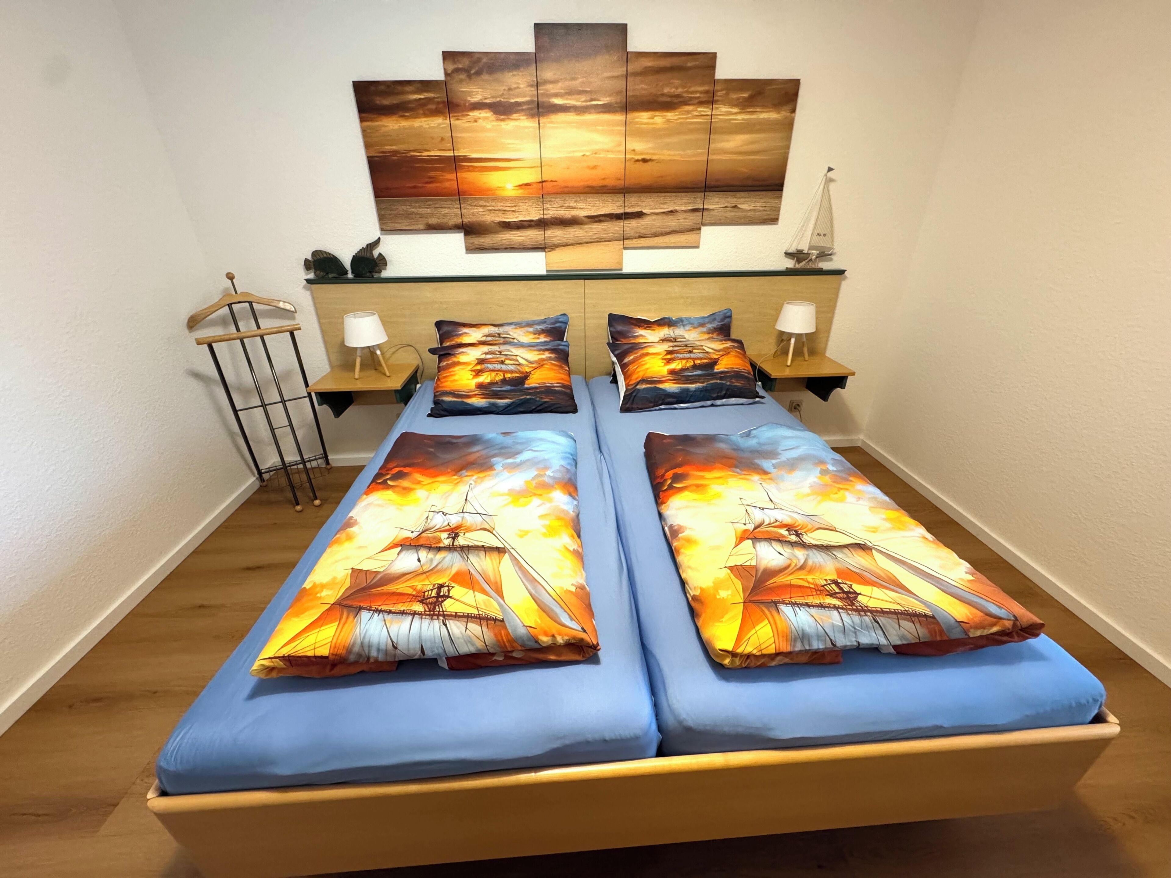 A double bed with matching blue bedding and pillows, a wall-mounted artwork depicting a sunset, and a small sailboat model on a shelf.