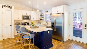 A modern kitchen with white cabinetry, a central island with chairs, stainless steel appliances, and a view through a glass door. - Summer STATus w/Optional Boat Rental (Mooresville)
