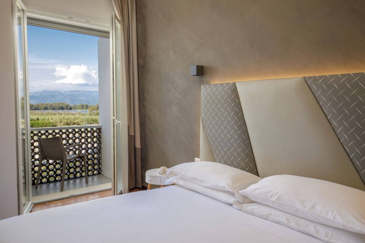 A hotel room with a bed, a nightstand, a chair, and a view of a balcony with a chair and a patterned wall.
