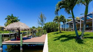 A dock with a thatched roof, a grassy area with palm trees, and a building with large windows. - Paradise Palms - Luxury Home - Bikes & Kayaks (Cape Coral)