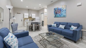 Una sala de estar moderna con un sofá azul, una mesa de centro de vidrio y una zona de cocina con armarios blancos y un microondas.