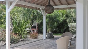 Een overdekt terras met een barbecue, zitplaatsen en weelderige tropische planten.