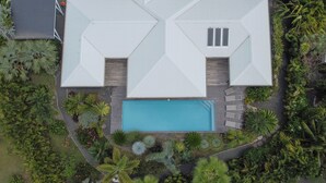 Een luchtendienst van een modern huis met een geometrisch dakontwerp, een zwembad en omliggende tropische begroeiing.