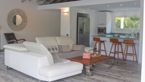 Een moderne woonkamer met een witte bank, een houten salontafel en een keukenhoek met een koelkast en barstoelen.