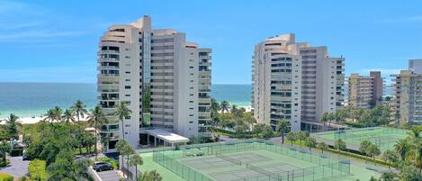 A high-rise hotel with multiple floors, a tennis court, and a view of the beach.