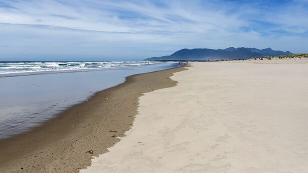 A beach with a wide sandy shore, a calm sea, and distant mountains under a clear sky. - Romantic A-Frame Beach Cottage, Blocks to the Beach, Deck, Fireplace and Firepit (Rockaway Beach)