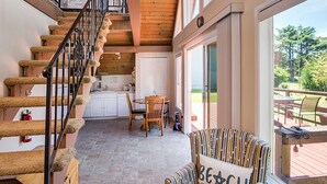 A wooden staircase with a black railing leading to an upper level. A striped armchair with a pillow that reads "BEACH" is placed near a large window overlooking a deck. - Romantic A-Frame Beach Cottage, Blocks to the Beach, Deck, Fireplace and Firepit (Rockaway Beach)