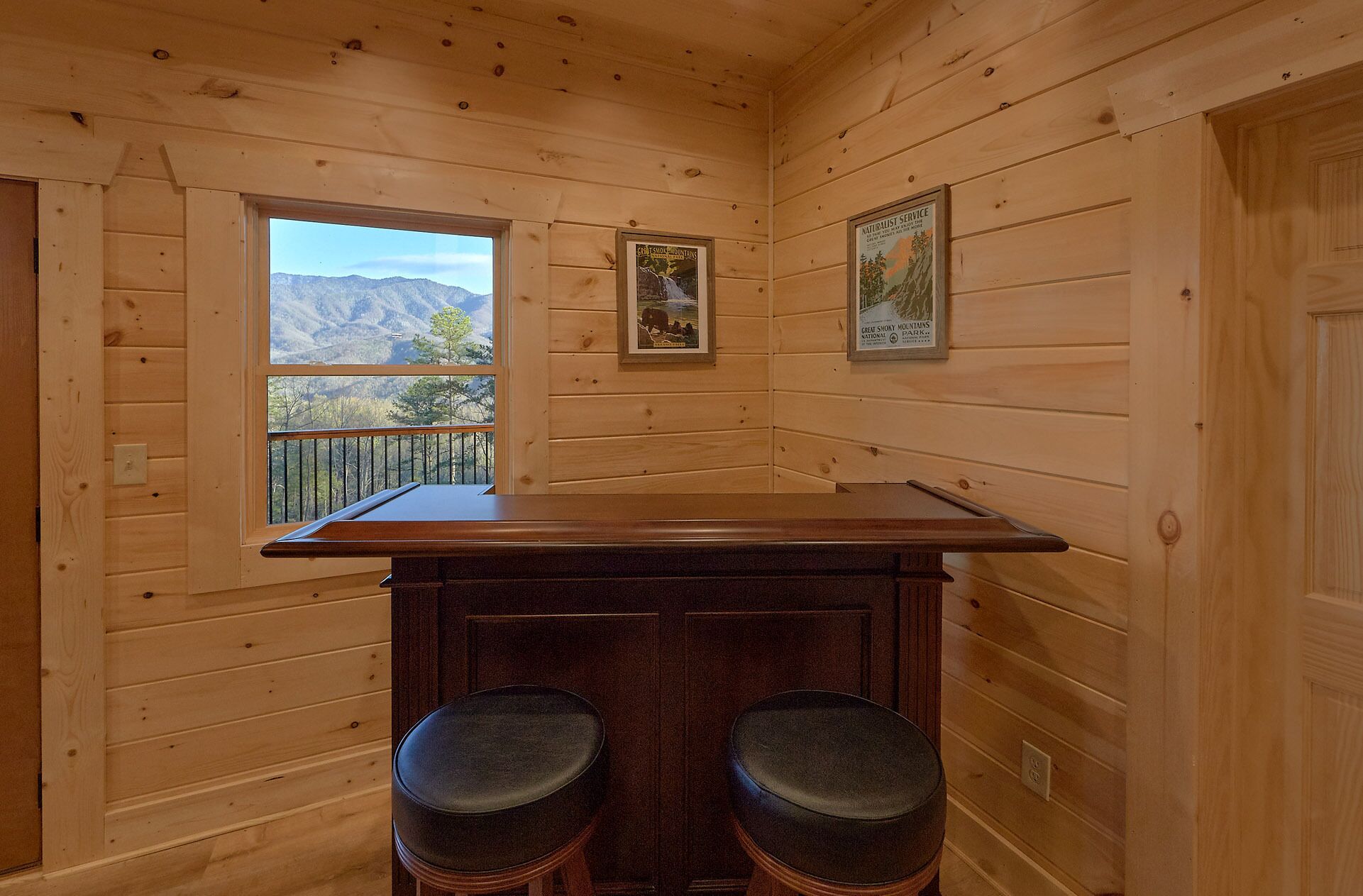 Habitación de cabaña de madera con ventana que ofrece vistas a la montaña, dos taburetes de madera y dos carteles enmarcados en la pared.