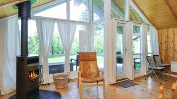 A cozy cabin interior with a wood-burning stove, large windows, a wooden ceiling, and a comfortable seating area.