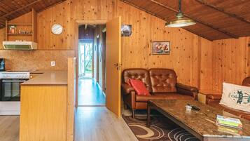 A cozy cabin interior with a wooden ceiling, a dining area with a table and chairs, a kitchen area with a stove and cabinets, and a hallway leading to a door.
