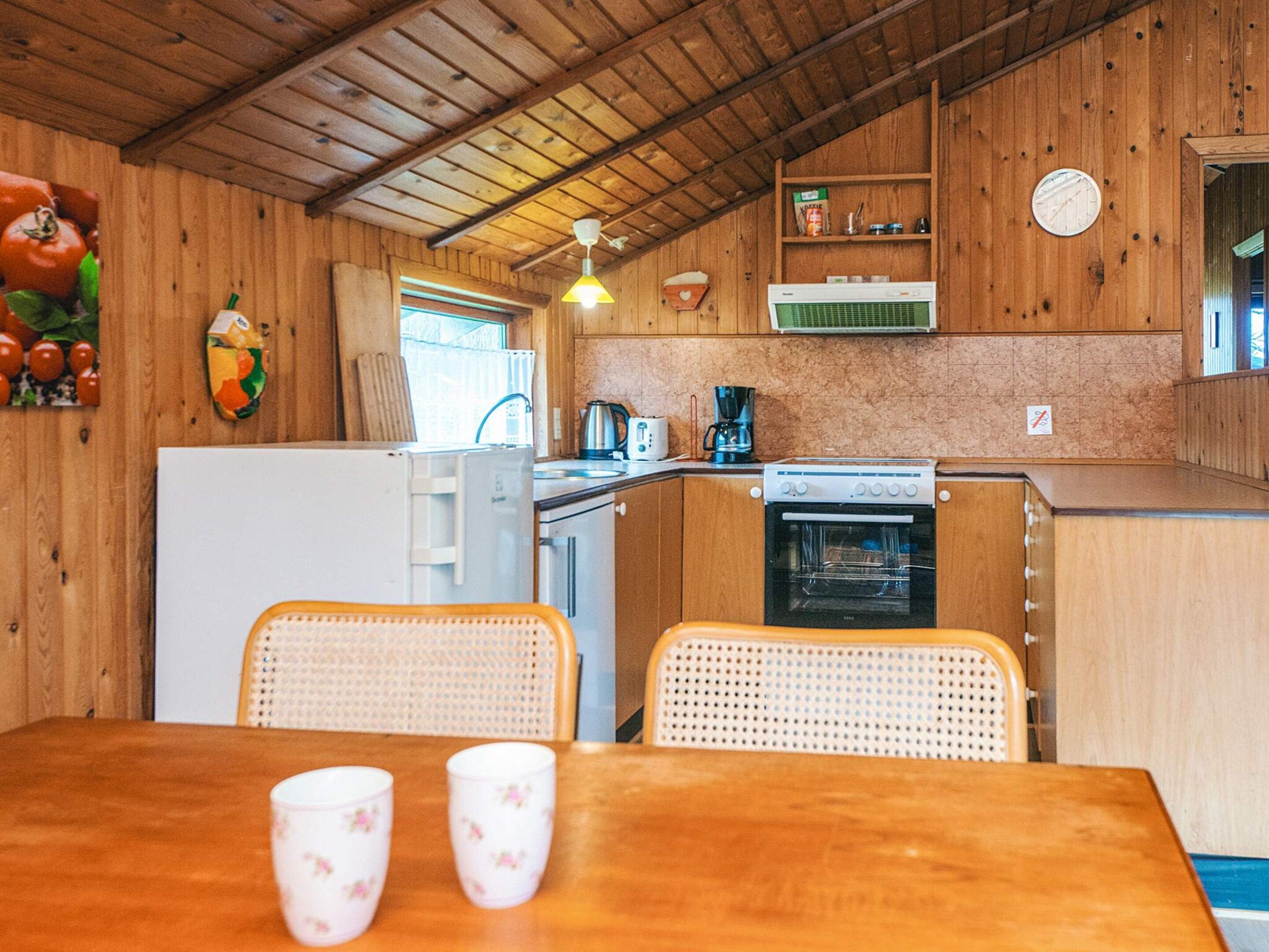 Pieni, käytännöllinen keittiö, jonka seinät ovat puuta. Keittiössä on jääkaappi, liesi ja pesuallas. Puupöydän ääressä on kaksi rottingista tuolia, ja pöydällä on kaksi kuppia.