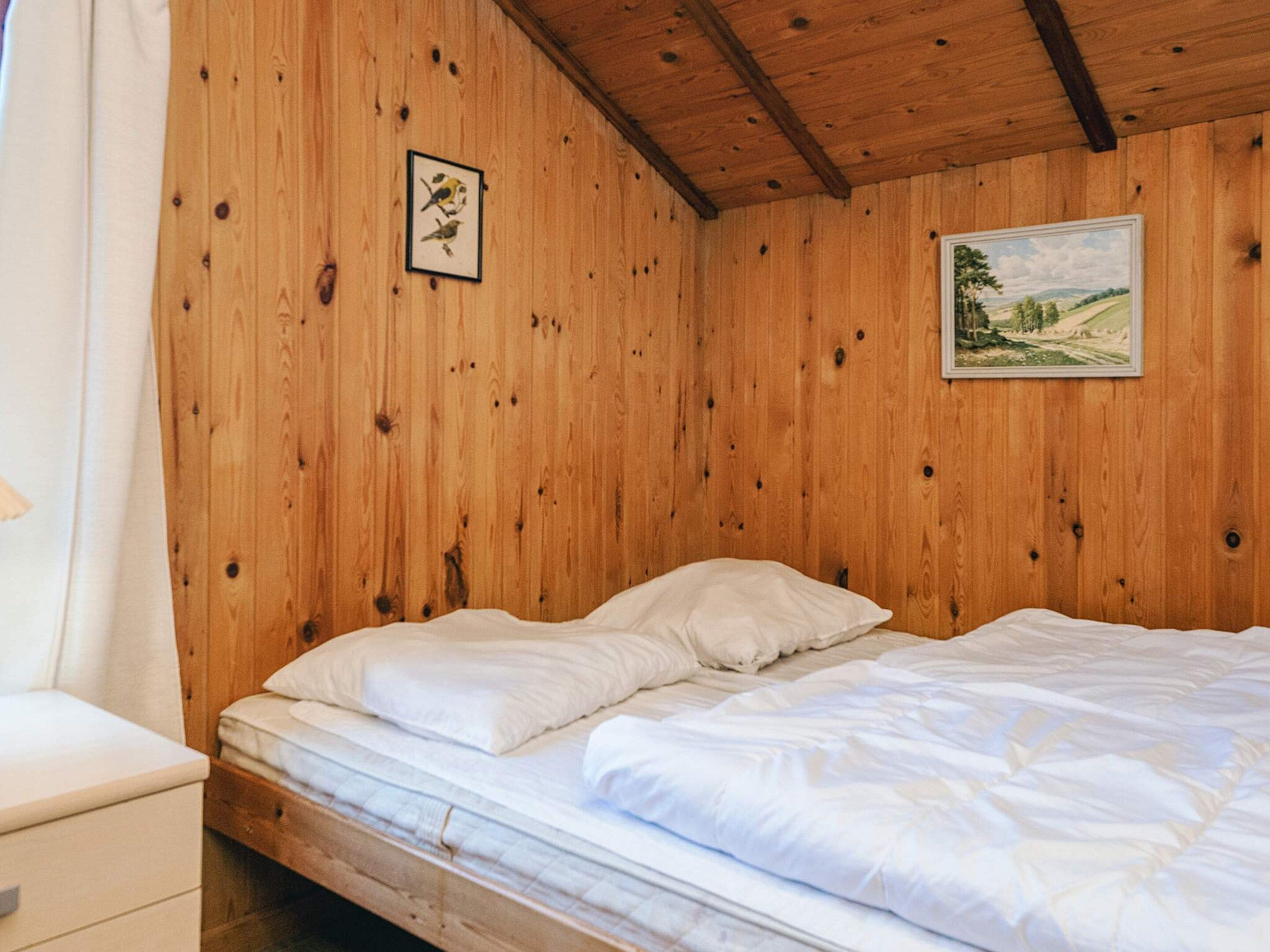 Una habitación de madera con una cama, una mesita de noche y dos cuadros en la pared.