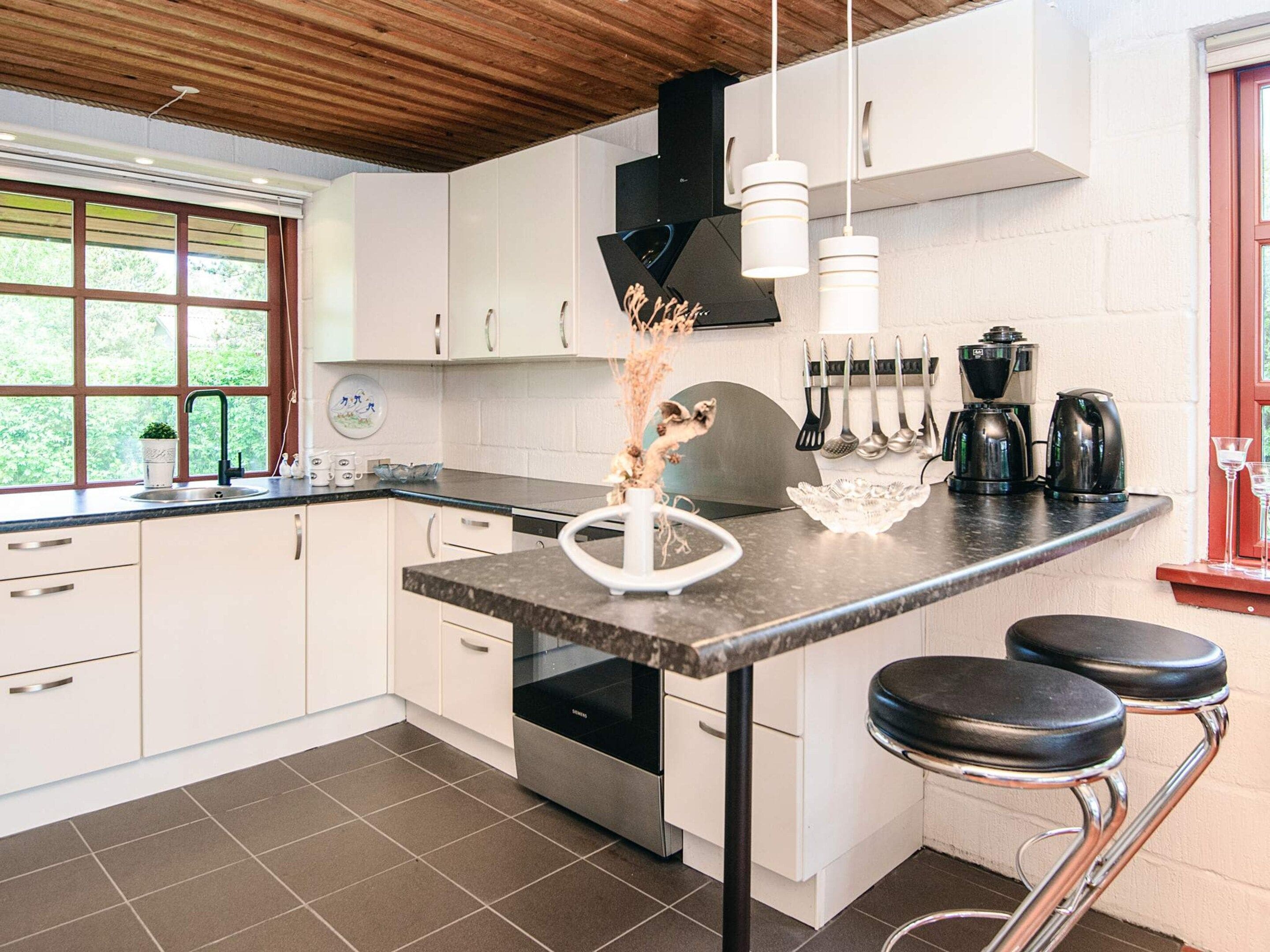A modern kitchen with white cabinets, a black countertop, and stainless steel appliances. There are two bar stools and a window with a view of greenery.