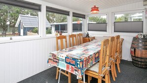 Un espace repas avec une longue table recouverte d’une nappe colorée, entourée de chaises en bois. Un luminaire suspendu rouge se trouve au-dessus de la table, et la pièce est dotée de murs lambrés blancs et de grandes fenêtres équipées de stores.