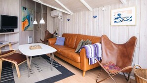 Un salon moderne avec un canapé en cuir marron, une table basse blanche et un fauteuil en osier. Un téléviseur à écran plat est fixé au mur, et un tableau est accroché au-dessus du canapé.
