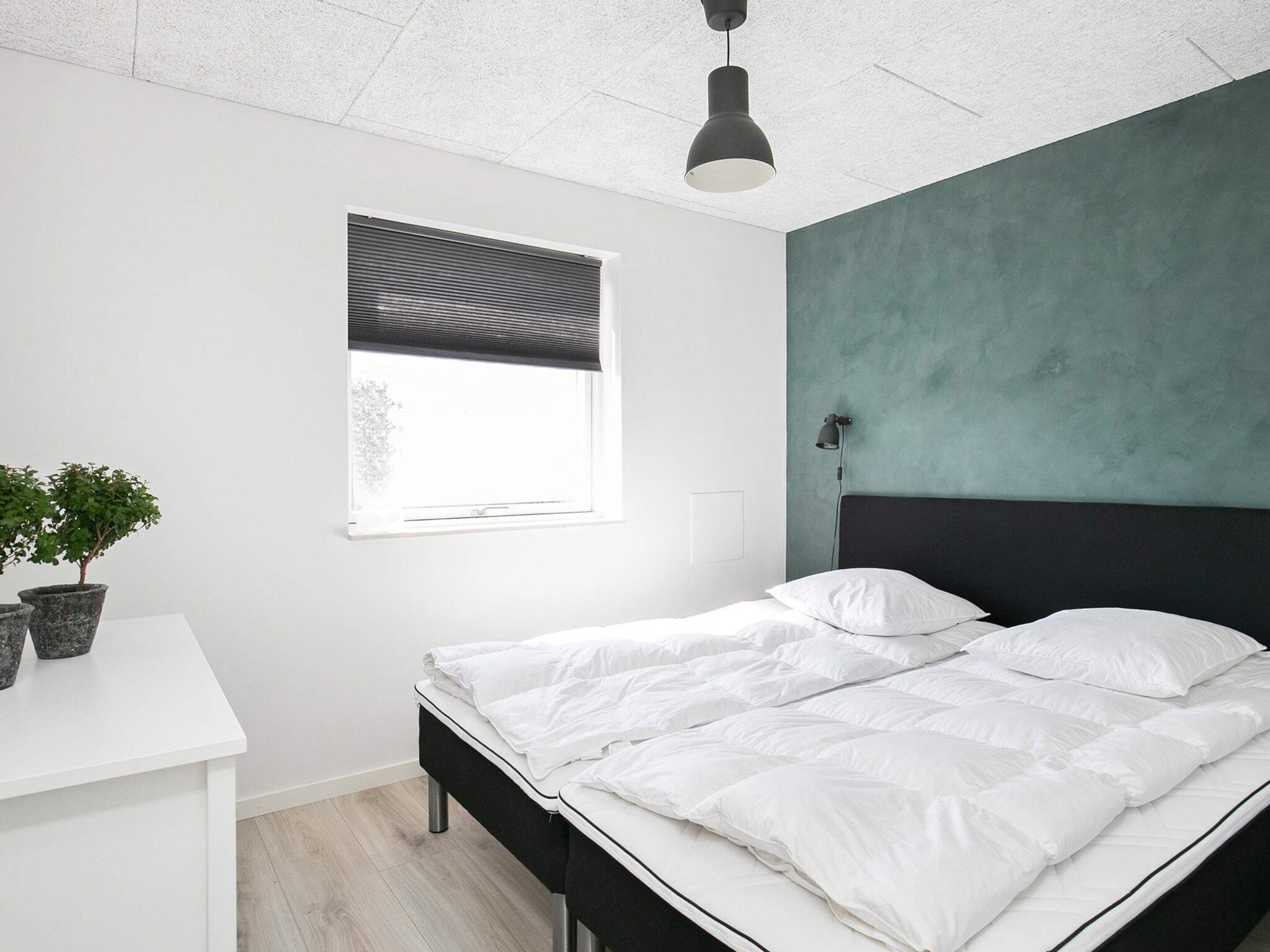 Un dormitorio con cama, escritorio, planta en maceta, ventana con persianas y una lámpara colgante.