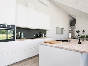 A modern kitchen with white cabinetry, a built-in oven, a coffee maker, and a wooden cutting board. - 4 Star Holiday Home in Vaeggerlose (Vaeggerlose)