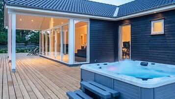 A modern outdoor hot tub area with a wooden deck, glass doors leading to a covered patio, and a dark-colored building.