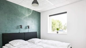 Une chambre moderne avec un lit, deux lampes de chevet, une fenêtre donnant sur des arbres et une voiture, et une petite table.