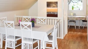 A dining area with a white table and chairs, a hanging light fixture, a window with patterned curtains, and a small kitchen area with a table and chairs. - 4 Star Holiday Home in Leirvik i Sogn (Hyllestad)