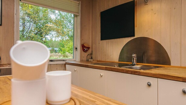 A kitchen with a wooden countertop, a window with a view of greenery, and a black screen mounted on the wall.