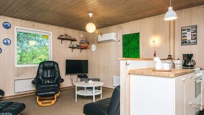 A compact living space with a black leather chair, a small round table, a TV, a kitchen area with a coffee maker, and a large window with a view of greenery.