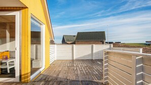 A wooden deck with a white fence, a yellow building, and a view of a house in the background. - 4 Person Holiday Home in Lemvig (Lemvig)