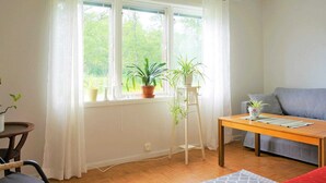 A room with a wooden floor, a grey sofa, a wooden table, and a red chair. There are potted plants on a windowsill and a floor lamp. - 4 Person Holiday Home in Gotene (Götene)