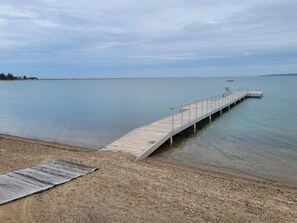 En träbrygga som sträcker sig ut i stilla vatten, med en sandstrand och ett metallgaller.