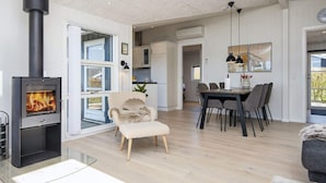 A modern living room with a wood-burning stove, a dining area with a table and chairs, and a cozy armchair. - Mod Holiday Home in Jutland near Sea (Lemvig)