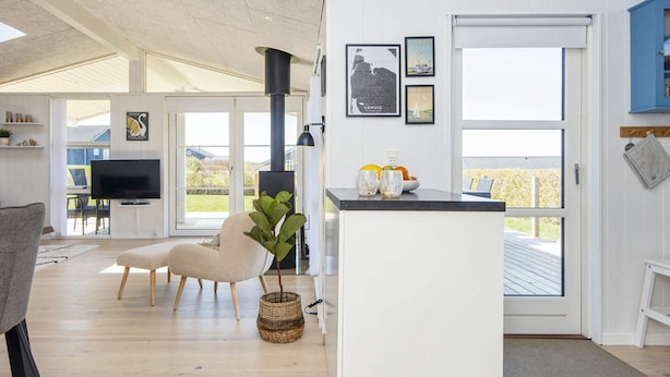 A modern living room with a TV, a comfortable chair, a small kitchen area with a counter, and a large window offering a view of the outdoors. - Mod Holiday Home in Jutland near Sea (Lemvig)