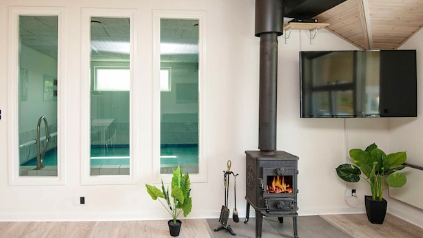 A modern interior with a wood-burning stove, large windows, and a flat-screen TV mounted on the wall. - Elegant Holiday Home in Jutland With Sauna (Horsens)