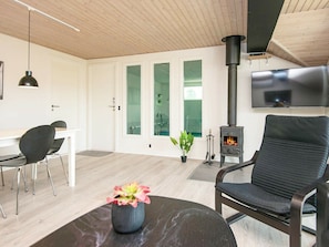 A modern living room with a black sofa, a dining table with chairs, a TV mounted on the wall, and a wood-burning stove.