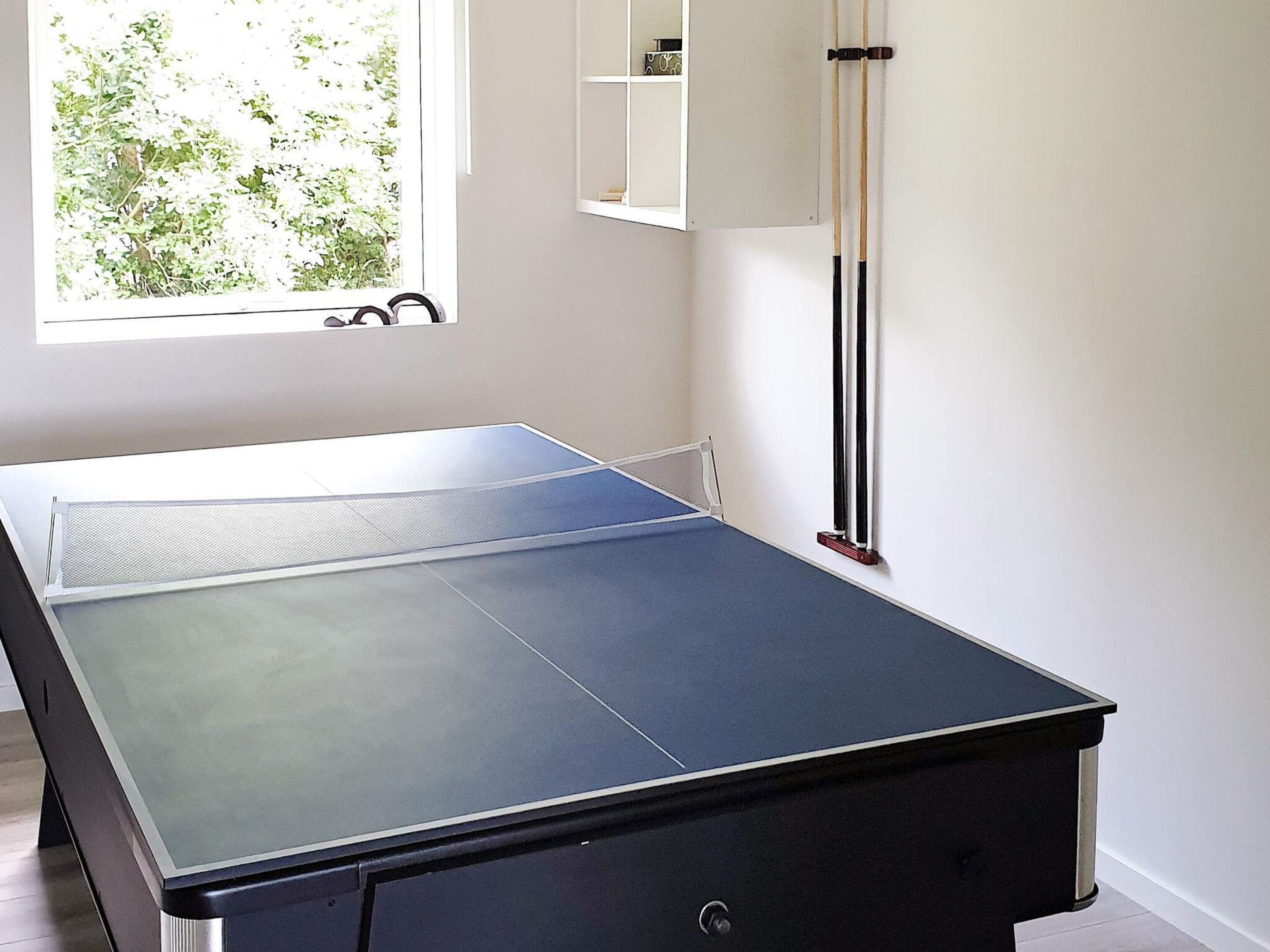 Et bordtennisbord i et rum med et vindue, en reol med genstande og et vægmonteret stativ.