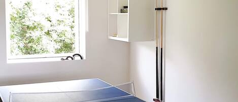 Une table de ping-pong dans une pièce avec une fenêtre, une étagère remplie d’objets et un porte-objets fixé au mur.
