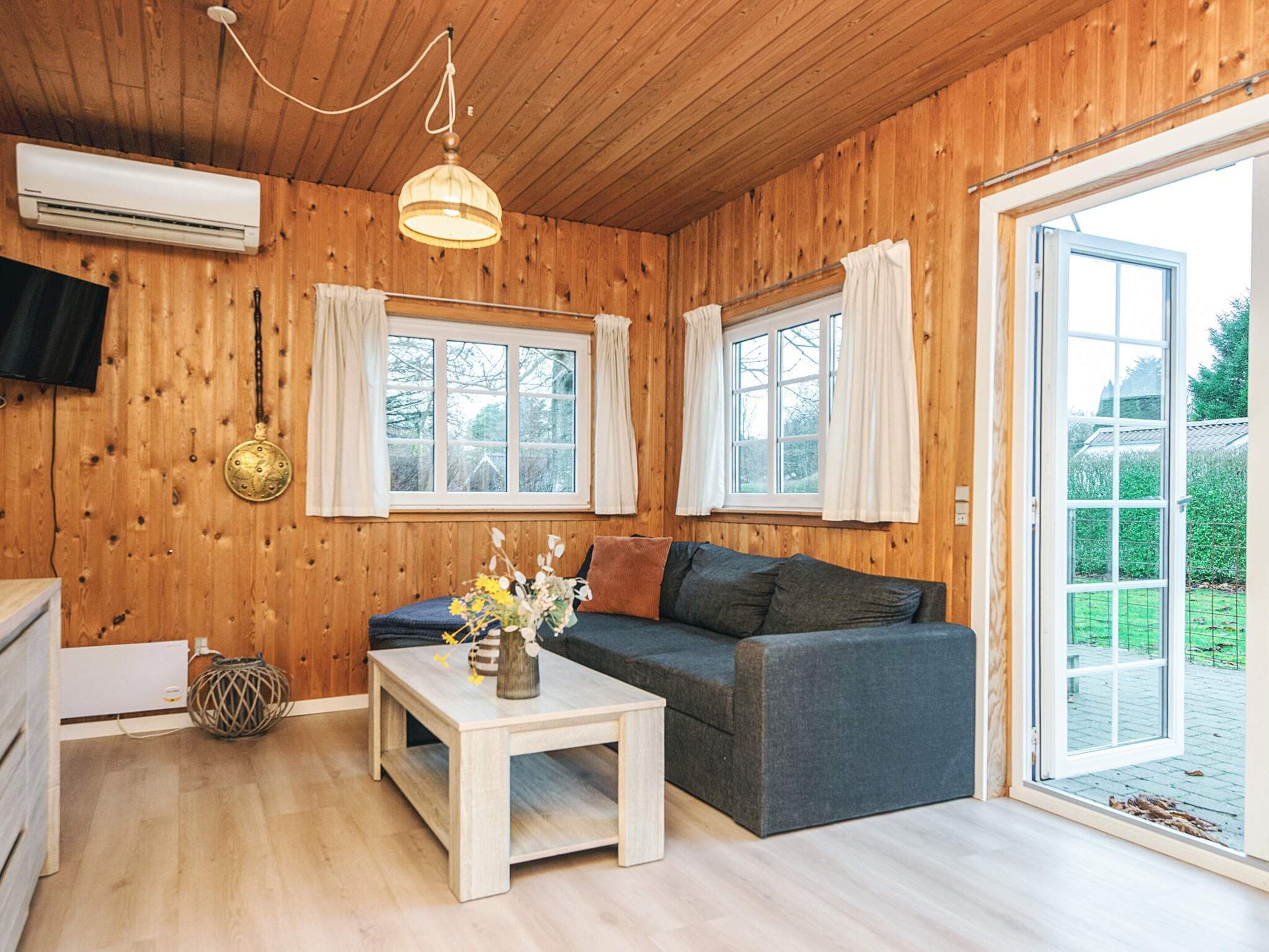 A cozy room with wooden walls, a grey sofa, a white table with a vase, and two windows with curtains.