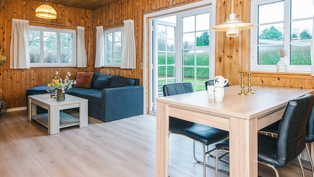 A cozy room with wooden walls, a sofa, a dining table with chairs, and large windows. - 4 Person Holiday Home in Ansager (Ansager)