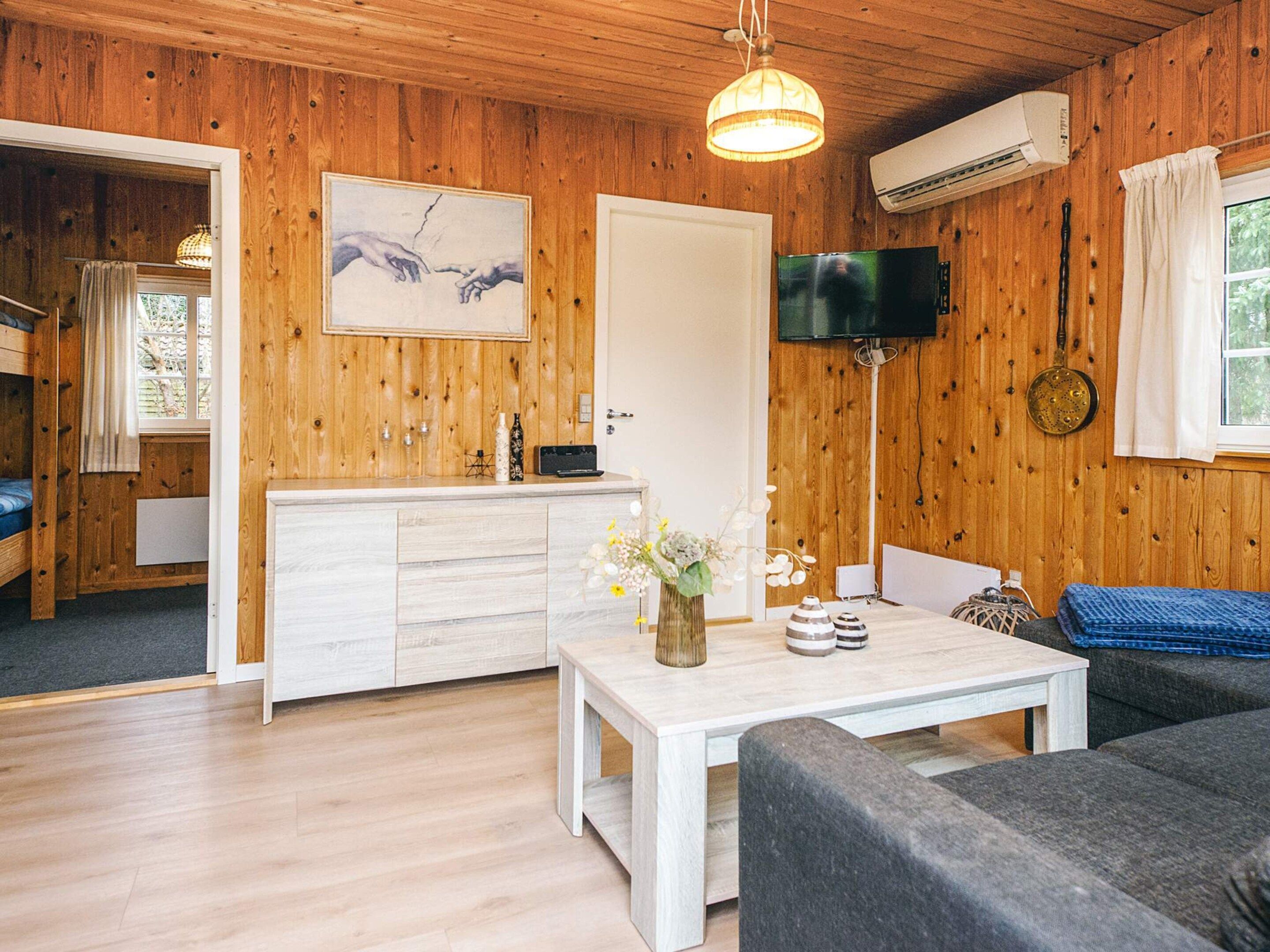 Ein gemütliches Blockhaus-Interieur mit Holzwänden, einem weißen Esstisch, einer Couch, einem an der Wand montierten Fernseher und einer Tür, die in einen anderen Raum führt.