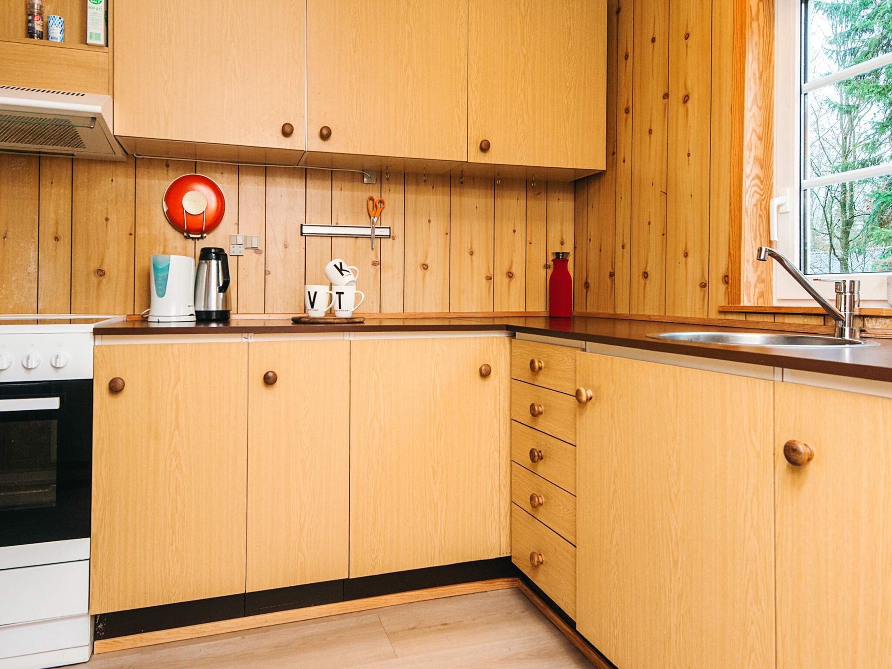 A kitchen with wooden cabinets, a stove, a sink, and a window with a view of trees.