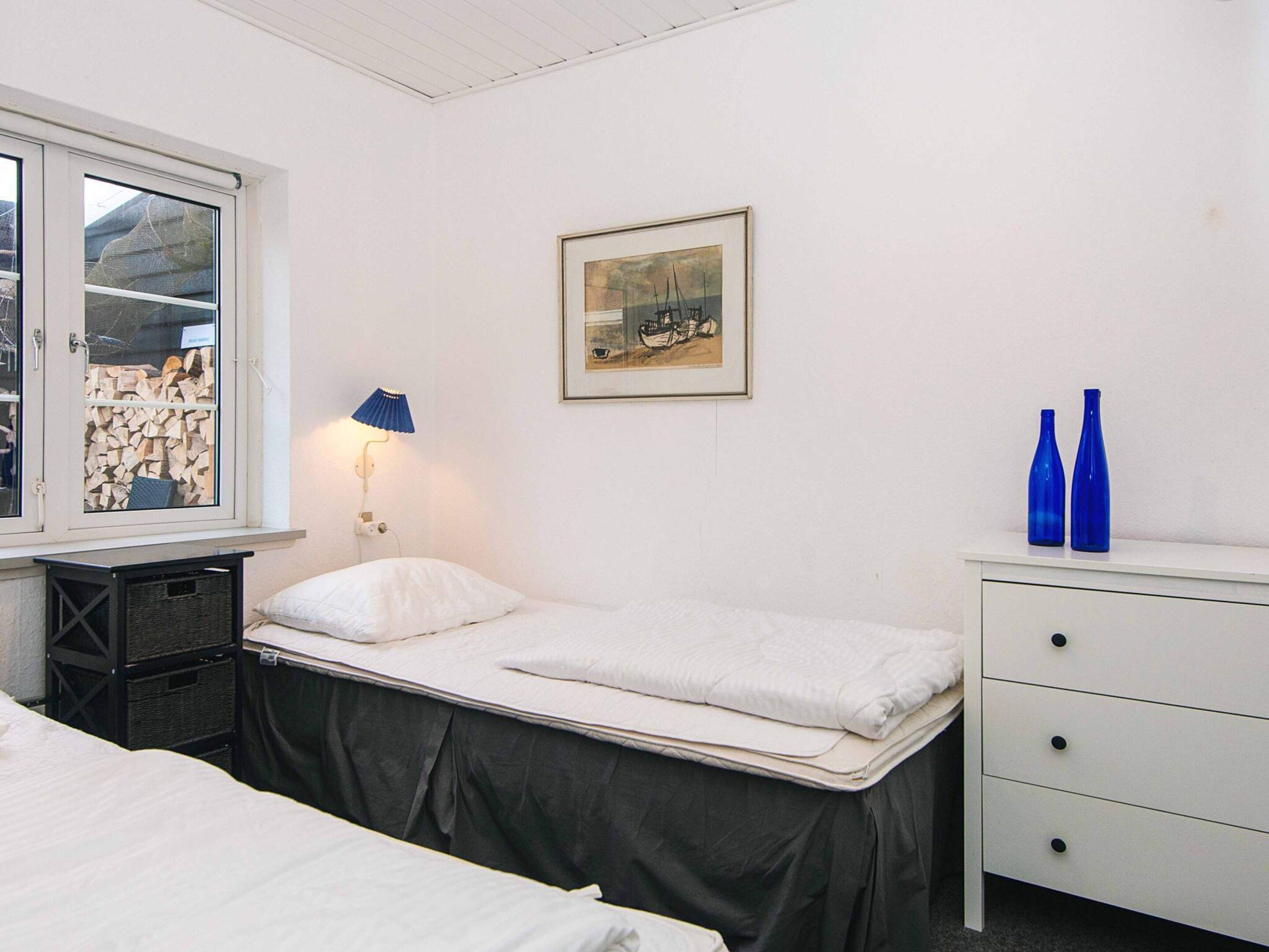 A room with two beds, a window with a view of stacked firewood, a wall-mounted lamp, a framed picture, and a dresser with two blue bottles.
