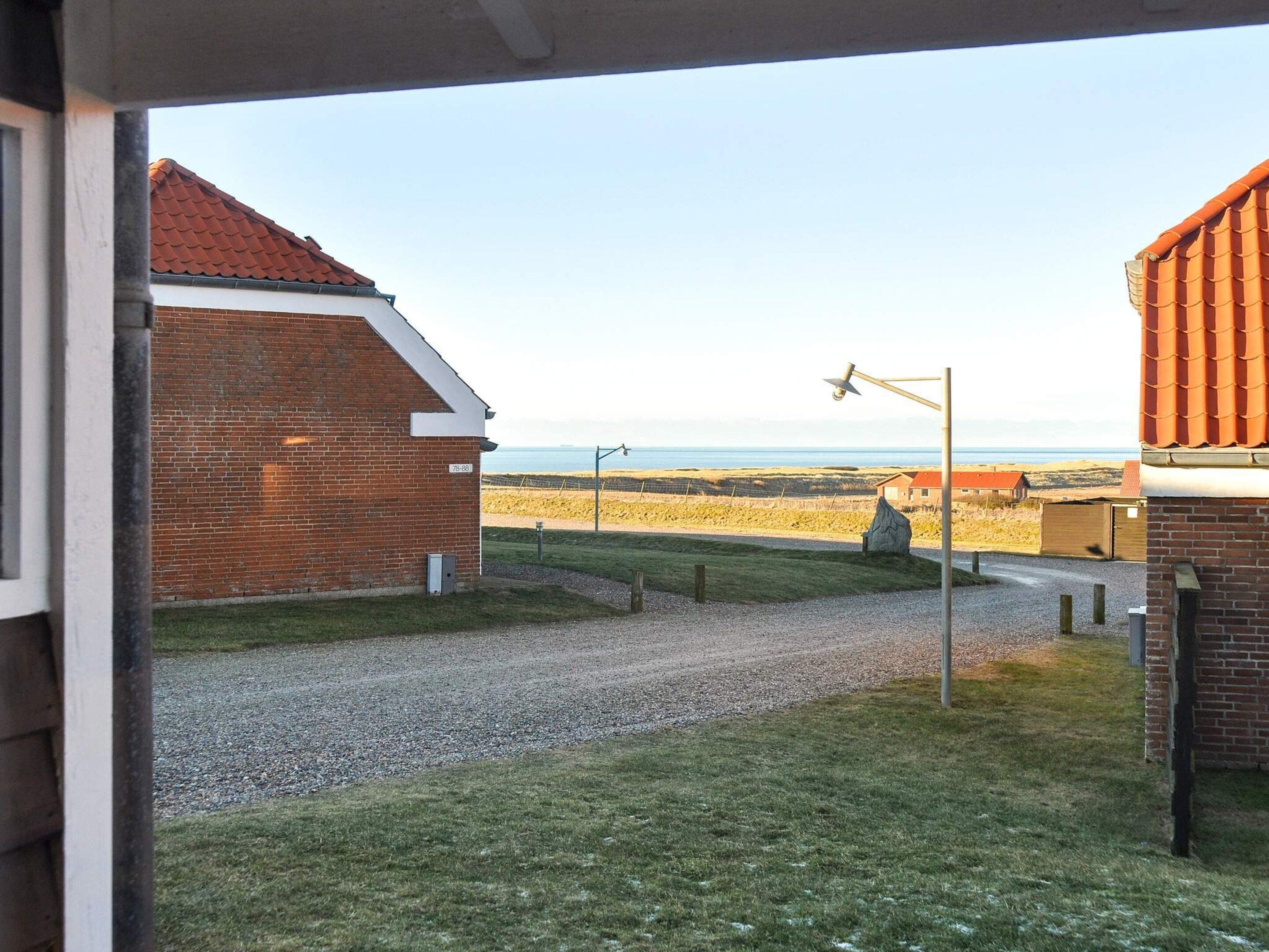 A view from a window overlooking a gravel driveway leading to a brick building with a red roof, a grassy field, and a distant view of the sea.
