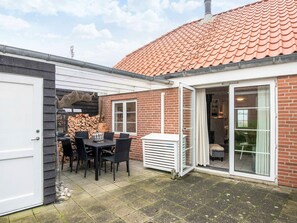Et murstenshus med tegltag, en overdækket terrasse med bord og stole, og en skydedør, der fører ind til et rum.