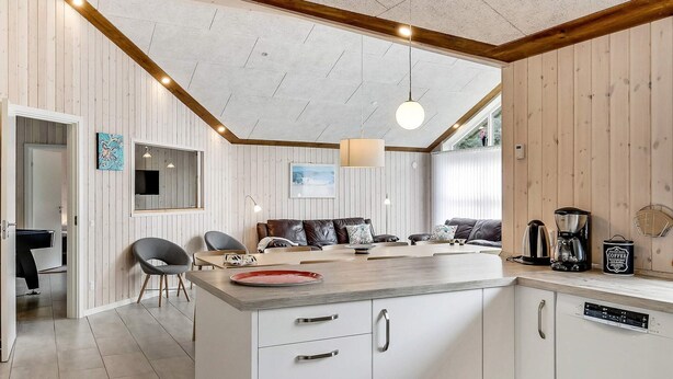 A modern kitchen with a central island, white cabinetry, and a wooden ceiling. There is a dining area with a sofa and chairs, and a coffee machine on the counter. - 4 Star Holiday Home in Oksbol (Oksbol)