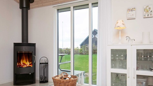 A modern living room with a wood-burning stove, a wicker basket with logs, a glass cabinet with glassware, and a view of a green lawn and a building outside. - Modernized Seaside Retreat-by Traum (Lokken)